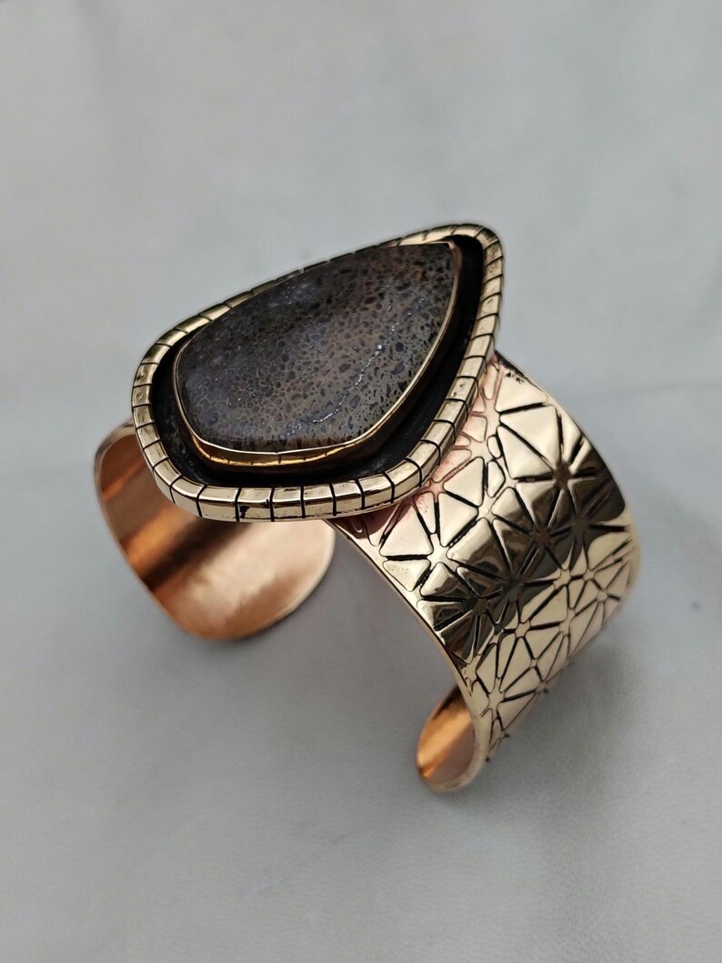 May include: A gold-coloured cuff bracelet featuring a large, teardrop-shaped stone. The stone is dark grey with a textured surface, set in a gold frame. The cuff has a geometric pattern.