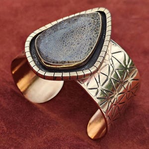 May include: A wide cuff bracelet featuring a large, teardrop-shaped stone. The stone is a mottled grey and brown colour, set in a gold-toned metal frame. The bracelet band has a geometric pattern and is also gold-toned.