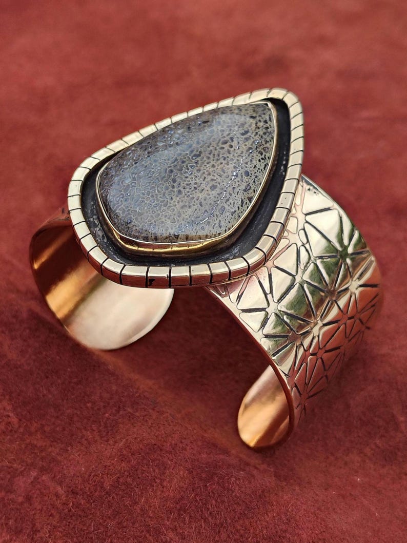 May include: A wide cuff bracelet featuring a large, teardrop-shaped stone. The stone is a mottled grey and brown colour, set in a gold-toned metal frame. The bracelet band has a geometric pattern and is also gold-toned.