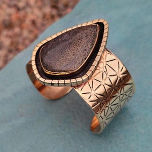May include: A gold-coloured cuff bracelet featuring a large, teardrop-shaped stone. The stone is dark grey with a speckled pattern, set in a black and gold frame. The cuff has a geometric starburst design.