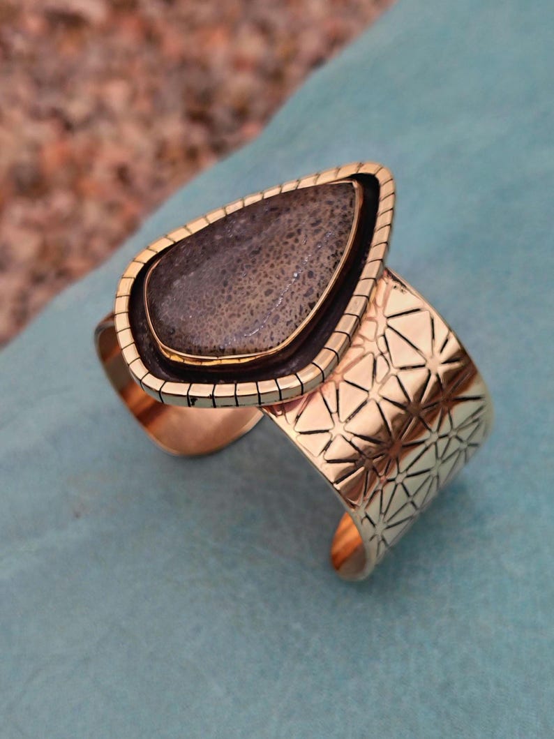 May include: A gold-coloured cuff bracelet featuring a large, teardrop-shaped stone. The stone is dark grey with a speckled pattern, set in a black and gold frame. The cuff has a geometric starburst design.