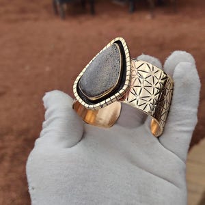 May include: Gold-coloured cuff bracelet with a large, teardrop-shaped stone displaying a grey and black pattern. The stone is set in a gold frame, and the cuff has a geometric design. A stylish piece of jewellery.