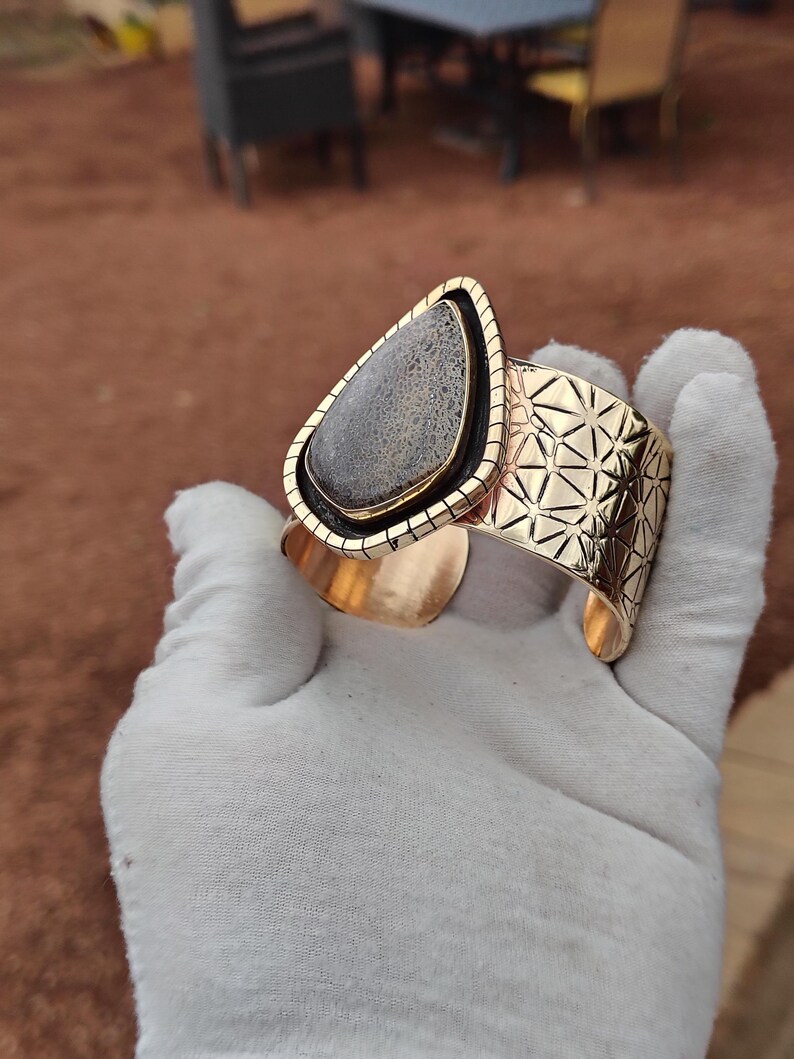 May include: Gold-coloured cuff bracelet with a large, teardrop-shaped stone displaying a grey and black pattern. The stone is set in a gold frame, and the cuff has a geometric design. A stylish piece of jewellery.