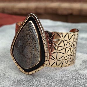 May include: A gold-coloured cuff bracelet featuring a large, teardrop-shaped stone. The stone is dark grey with a textured pattern, set in a black border. The cuff has a geometric starburst design.