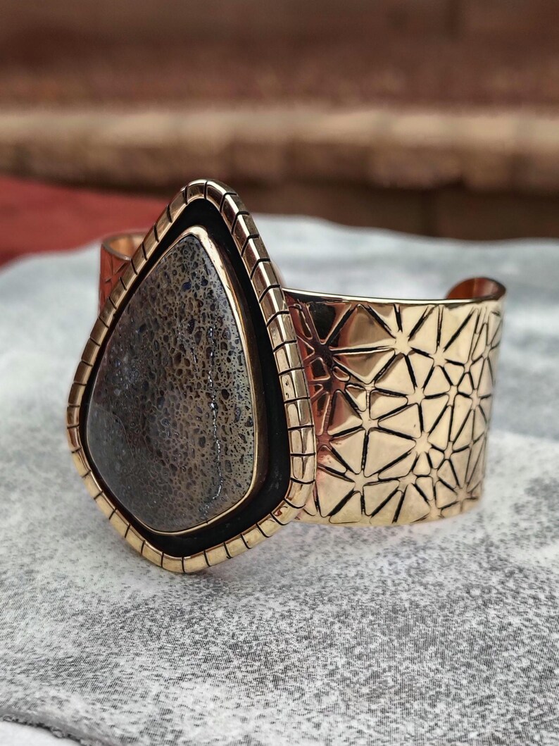 May include: A gold-coloured cuff bracelet featuring a large, teardrop-shaped stone. The stone is dark grey with a textured pattern, set in a black border. The cuff has a geometric starburst design.