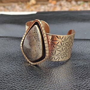 May include: A gold-coloured cuff bracelet featuring a large, teardrop-shaped stone. The stone is a mix of beige and brown with a textured appearance. The cuff has geometric patterns and a black border around the stone.