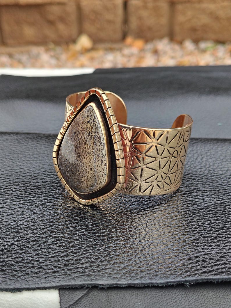 May include: A gold-coloured cuff bracelet featuring a large, teardrop-shaped stone. The stone is a mix of beige and brown with a textured appearance. The cuff has geometric patterns and a black border around the stone.
