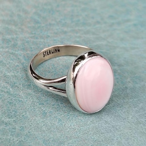 May include: A sterling silver ring featuring a smooth, oval-shaped light pink cabochon stone. The metal band has a split design and the word STERLING is clearly stamped on the inner surface of the band, indicating the metal composition of the ring.