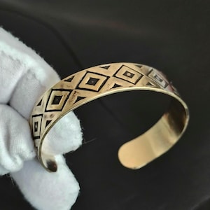 May include: A gold-toned metal cuff bracelet featuring a repeating pattern of black-filled diamonds and squares etched along the outer surface. The bracelet has an open-ended design with rounded terminals and a polished, reflective finish.