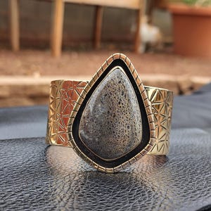 May include: A wide gold-coloured cuff bracelet featuring a large, teardrop-shaped stone. The stone is grey with a textured pattern, framed by black and gold. The cuff has a geometric design.