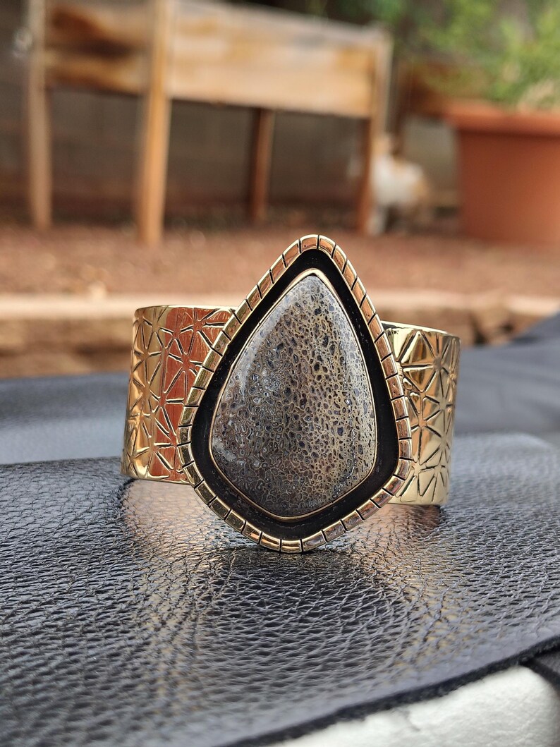 May include: A wide gold-coloured cuff bracelet featuring a large, teardrop-shaped stone. The stone is grey with a textured pattern, framed by black and gold. The cuff has a geometric design.