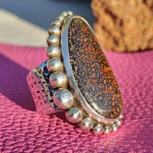 May include: A silver ring with a large, oval-shaped stone. The stone has a brown and orange pattern. The ring has a textured band and is decorated with small silver beads around the stone.