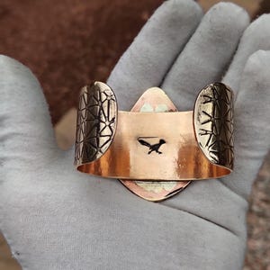 May include: Copper cuff bracelet featuring a roadrunner silhouette. The bracelet has geometric patterned ends and a central, diamond-shaped design. The cuff is held open, showing the inside with the roadrunner image.
