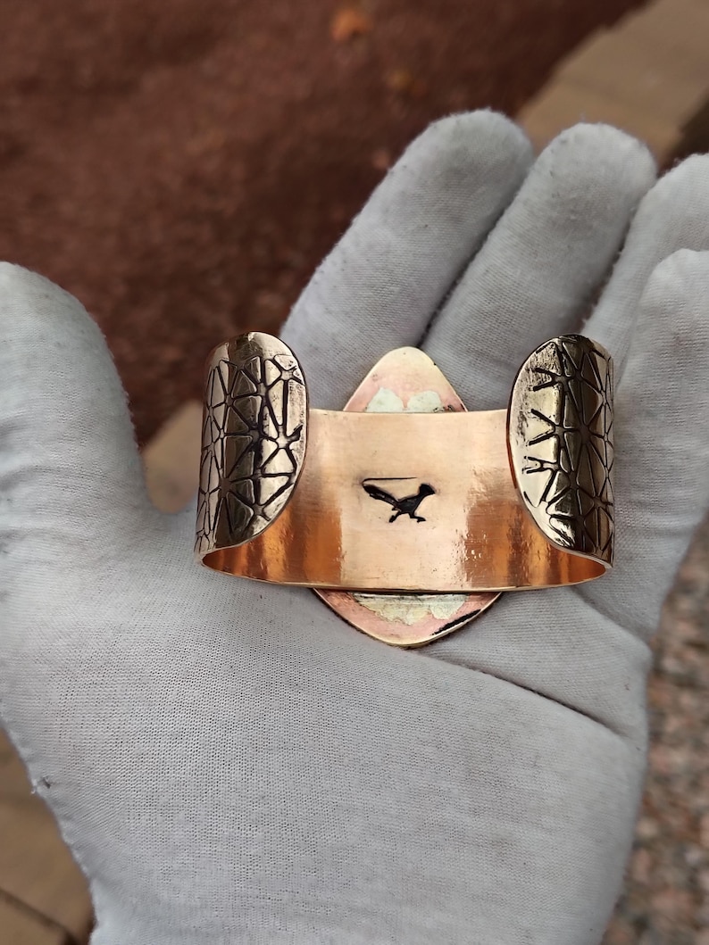May include: Copper cuff bracelet featuring a roadrunner silhouette. The bracelet has geometric patterned ends and a central, diamond-shaped design. The cuff is held open, showing the inside with the roadrunner image.