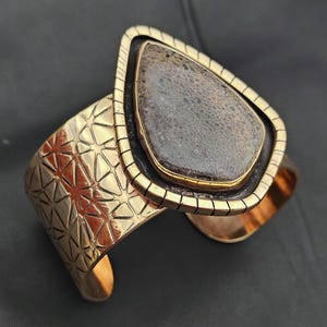 May include: A gold-coloured cuff bracelet featuring a large, teardrop-shaped stone. The stone is a mottled grey and brown colour, set in a black and gold frame. The cuff has a geometric pattern.