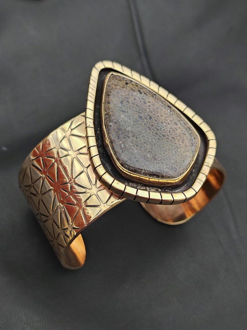 May include: A gold-coloured cuff bracelet featuring a large, teardrop-shaped stone. The stone is a mottled grey and brown colour, set in a black and gold frame. The cuff has a geometric pattern.