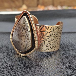 May include: A gold-coloured cuff bracelet with a large, teardrop-shaped stone. The stone is a mix of brown and grey hues, set in a gold frame. The cuff features a geometric pattern on the band.
