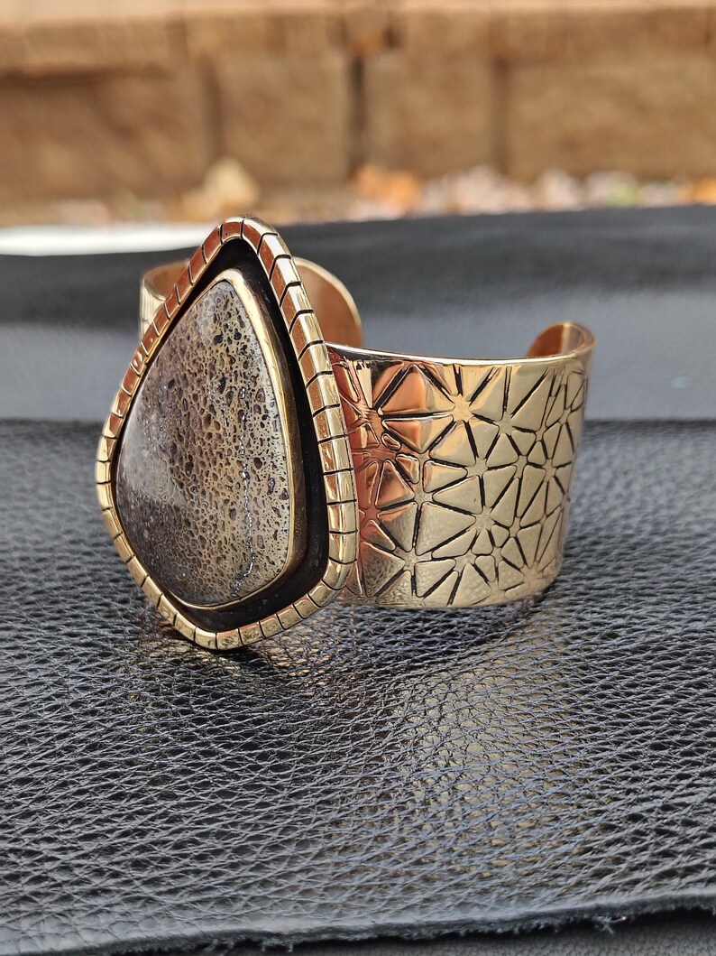 May include: A gold-coloured cuff bracelet with a large, teardrop-shaped stone. The stone is a mix of brown and grey hues, set in a gold frame. The cuff features a geometric pattern on the band.