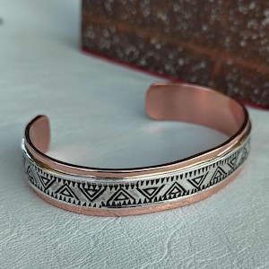 May include: A copper cuff bracelet featuring a central silver-toned band with a repeating black geometric triangle pattern. The bracelet has an open design, showcasing the metallic copper finish on the edges and the interior surface of the band.