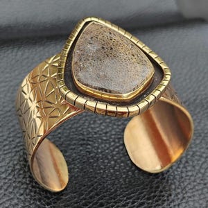 May include: A gold-coloured cuff bracelet featuring a geometric pattern and a triangular, fossil-like stone centrepiece. The stone is framed in gold and set on a dark brown backing. The bracelet has an open design, with a visible opening.
