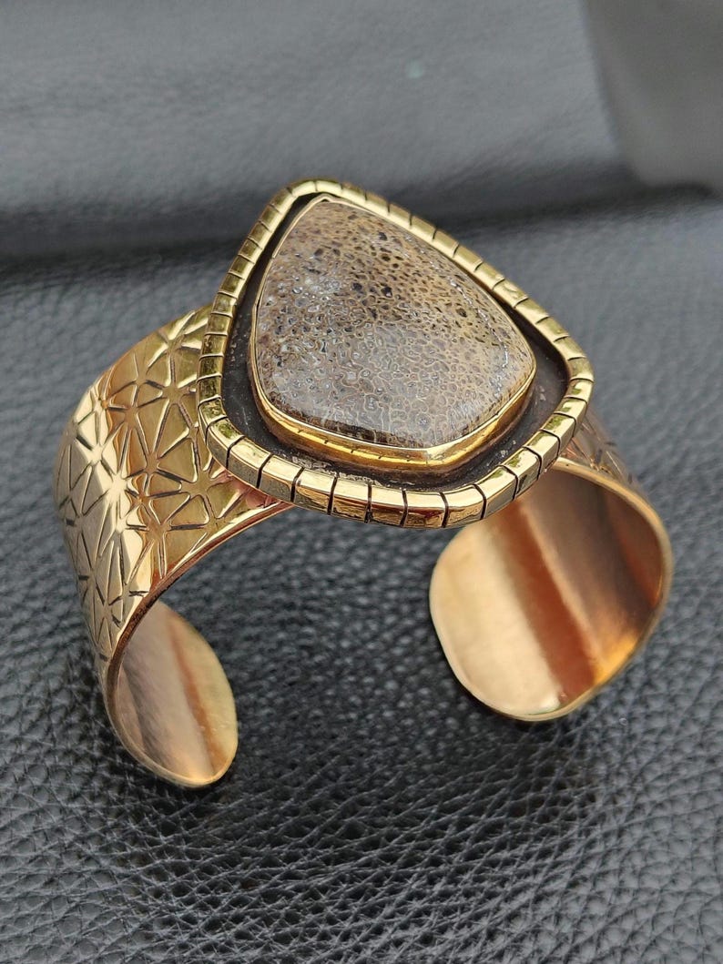 May include: A gold-coloured cuff bracelet featuring a geometric pattern and a triangular, fossil-like stone centrepiece. The stone is framed in gold and set on a dark brown backing. The bracelet has an open design, with a visible opening.