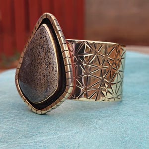 May include: A wide, gold-coloured cuff bracelet with a large, teardrop-shaped stone. The stone is a mix of brown and grey tones, set in a dark border. The cuff features a geometric pattern and a textured edge.