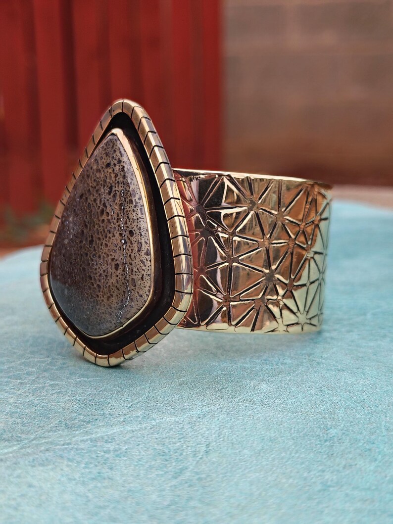 May include: A wide, gold-coloured cuff bracelet with a large, teardrop-shaped stone. The stone is a mix of brown and grey tones, set in a dark border. The cuff features a geometric pattern and a textured edge.