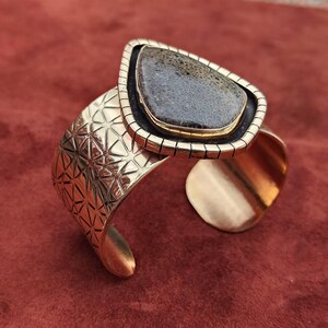 May include: A gold-coloured cuff bracelet featuring a large, teardrop-shaped stone. The stone is a dark grey colour with a textured pattern. The bracelet has a geometric design on the band and a contrasting black border around the stone.