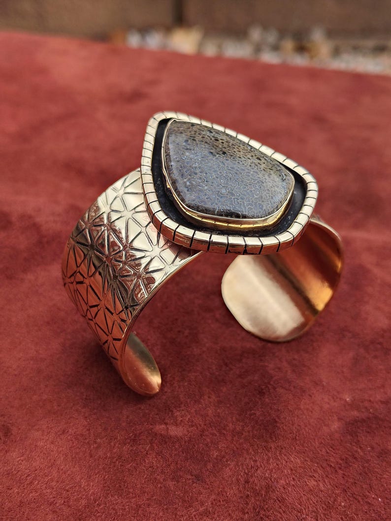 May include: A gold-coloured cuff bracelet featuring a large, teardrop-shaped stone. The stone is a dark grey colour with a textured pattern. The bracelet has a geometric design on the band and a contrasting black border around the stone.