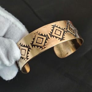 May include: A gold-toned metal cuff bracelet featuring a repeating pattern of etched diamonds surrounded by sunburst-like rays. The surface of the bracelet has a polished finish with darkened recessed areas that highlight the geometric design.