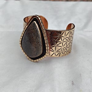 May include: A gold-coloured cuff bracelet featuring a large, teardrop-shaped stone. The stone is dark grey with a speckled pattern. The bracelet has a geometric design on the band and a textured frame around the stone.