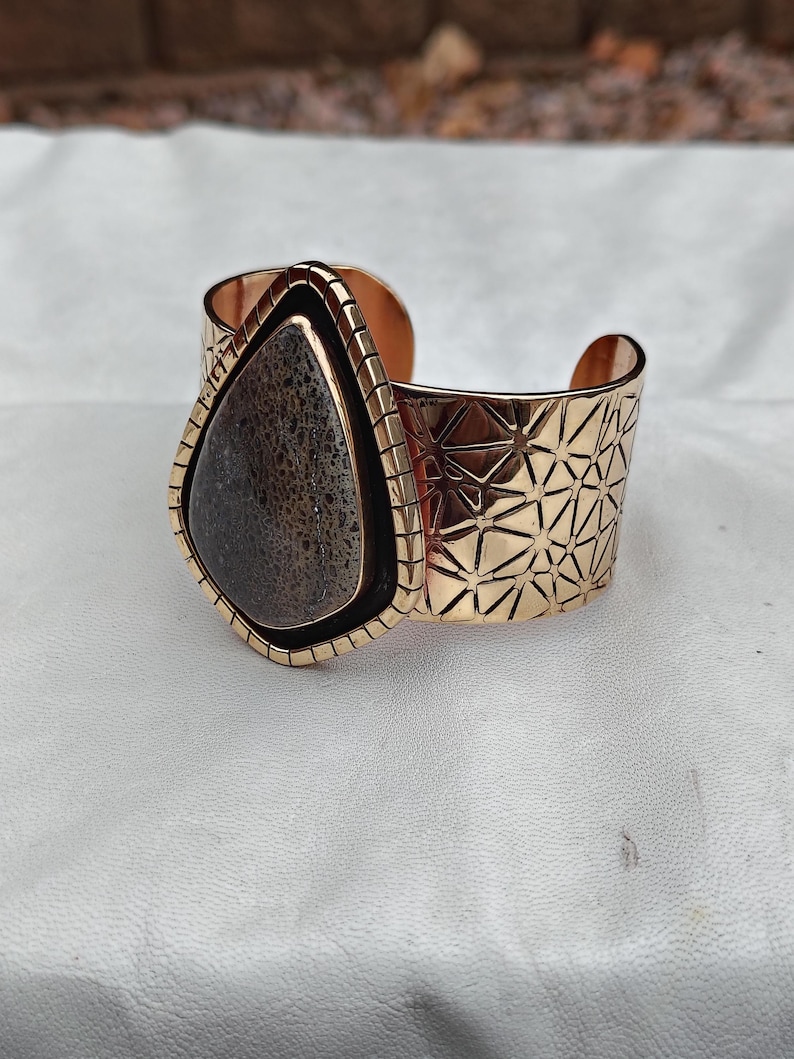 May include: A gold-coloured cuff bracelet featuring a large, teardrop-shaped stone. The stone is dark grey with a speckled pattern. The bracelet has a geometric design on the band and a textured frame around the stone.