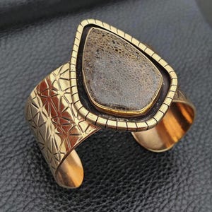 May include: A gold-coloured cuff bracelet featuring a large, teardrop-shaped stone. The stone is a mottled brown colour, set within a black and gold frame. The cuff has a geometric starburst pattern and a textured design.