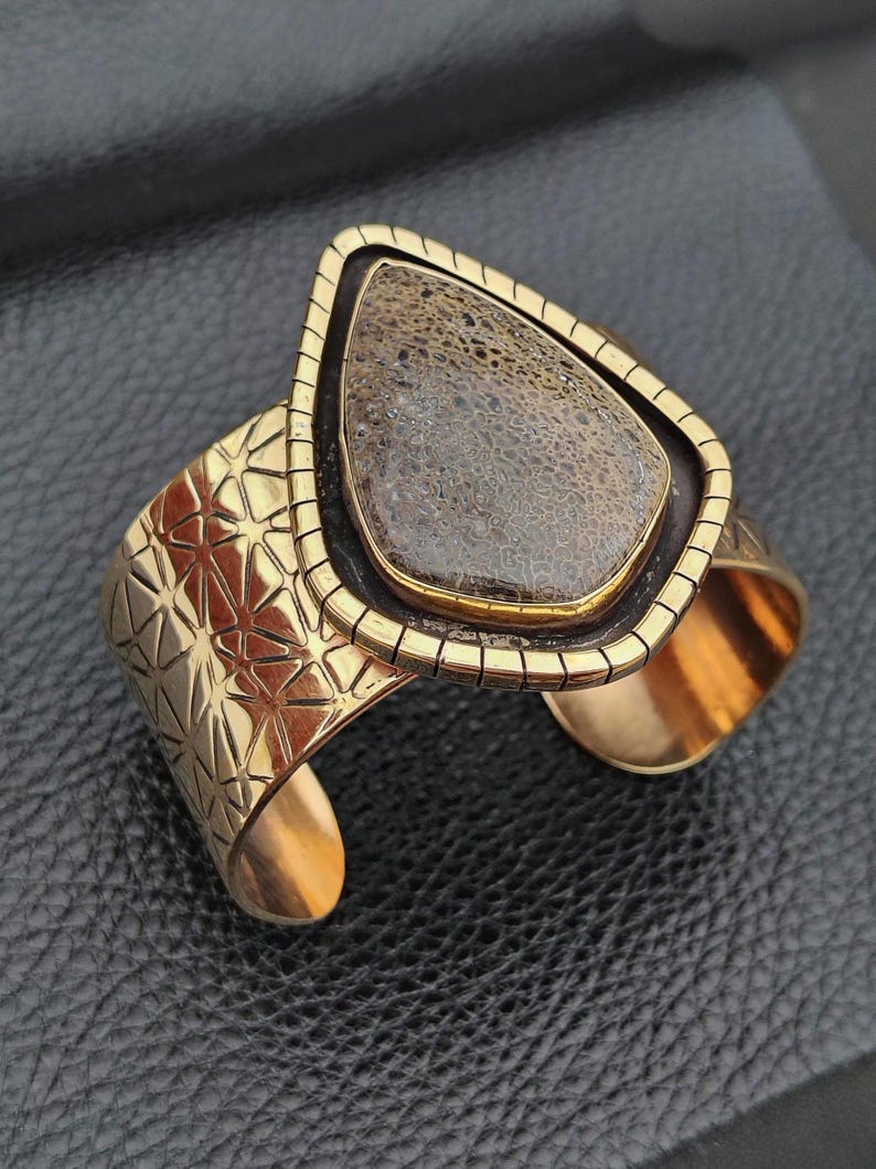May include: A gold-coloured cuff bracelet featuring a large, teardrop-shaped stone. The stone is a mottled brown colour, set within a black and gold frame. The cuff has a geometric starburst pattern and a textured design.