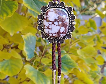May include: A bolo tie with a brown braided leather cord and a decorative pendant. The pendant has a silver-colored frame with a central stone featuring white and brown patterns. The tie's tips are gold-colored.