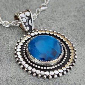 May include: A round pendant necklace featuring a central polished blue cabochon stone set in a silver frame. The frame has a dark oxidized finish with a decorative border of small silver spheres. It hangs from a silver rope chain necklace.
