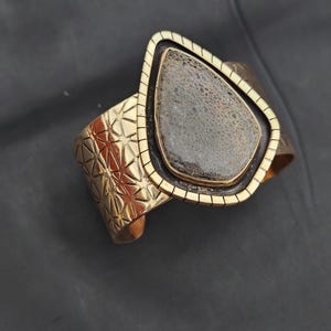 May include: A gold-coloured cuff bracelet featuring a geometric design and a large, teardrop-shaped stone. The stone is a mix of grey and brown with a textured surface. The bracelet has a rectangular frame around the stone.