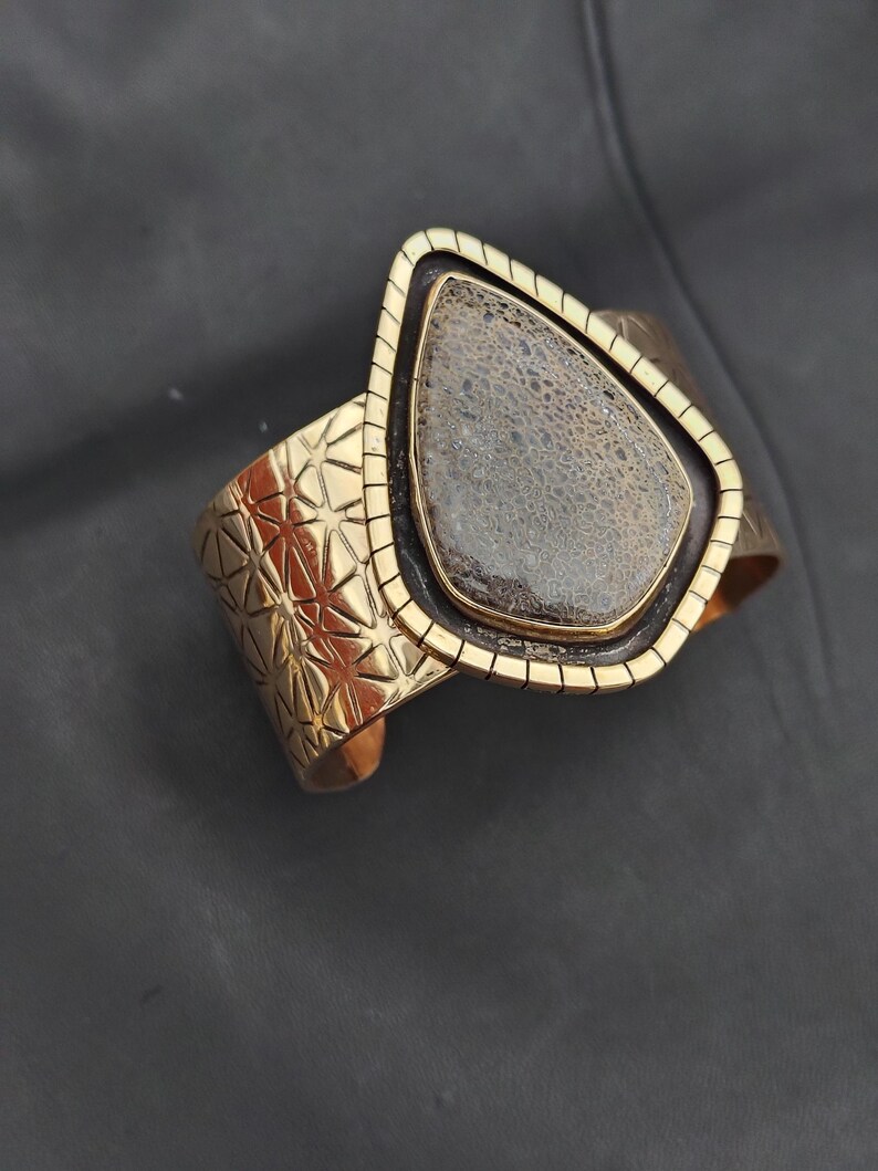 May include: A gold-coloured cuff bracelet featuring a geometric design and a large, teardrop-shaped stone. The stone is a mix of grey and brown with a textured surface. The bracelet has a rectangular frame around the stone.