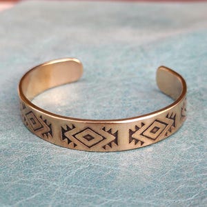 May include: A gold-colored cuff bracelet with an open design. The bracelet features a repeating geometric pattern of diamonds and triangles etched around the band. The bracelet is displayed on a light blue surface.