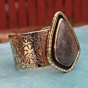 May include: A gold-coloured cuff bracelet with a geometric pattern and a large, teardrop-shaped stone. The stone is dark grey with a textured surface, set in a gold frame. The bracelet is on a light blue surface.