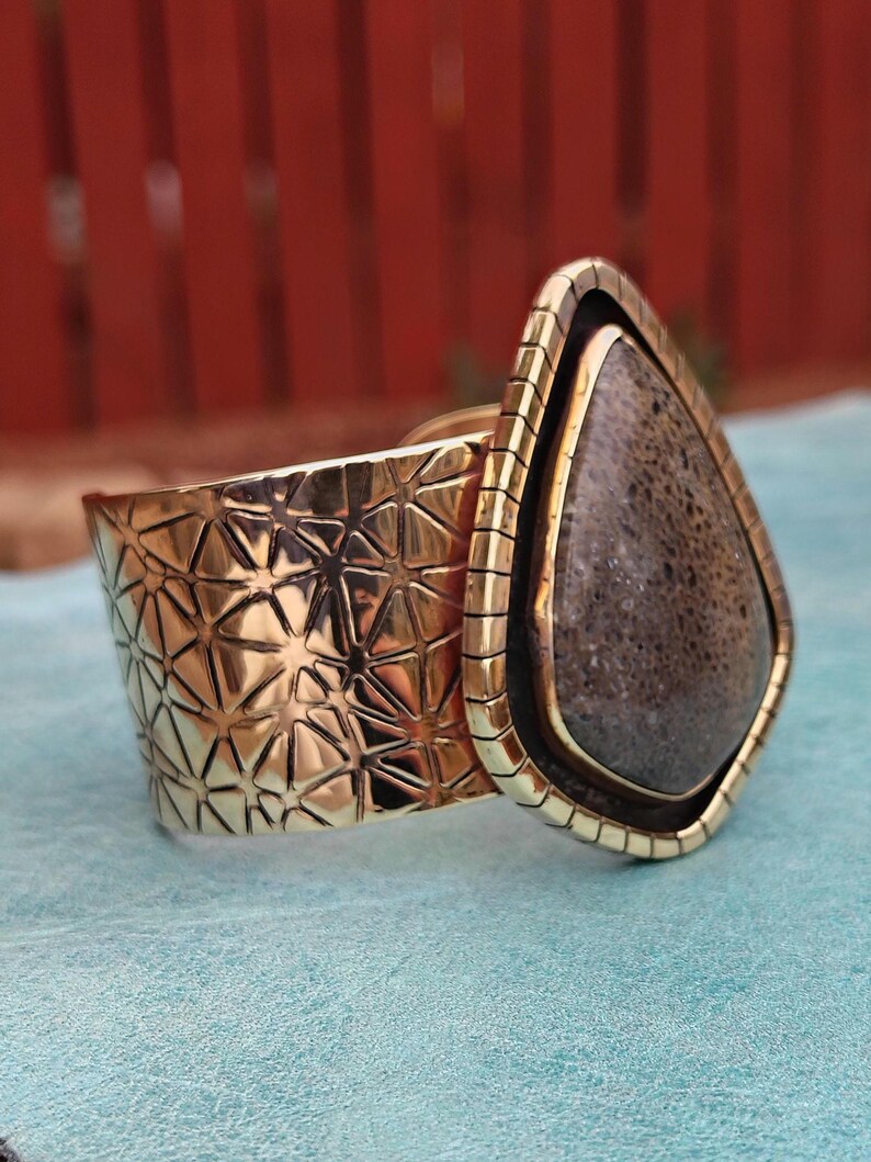 May include: A gold-coloured cuff bracelet with a geometric pattern and a large, teardrop-shaped stone. The stone is dark grey with a textured surface, set in a gold frame. The bracelet is on a light blue surface.