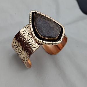 May include: A gold-coloured cuff bracelet featuring a geometric pattern and a teardrop-shaped, dark grey stone. The stone is set in a black and gold frame. The bracelet has a textured surface.