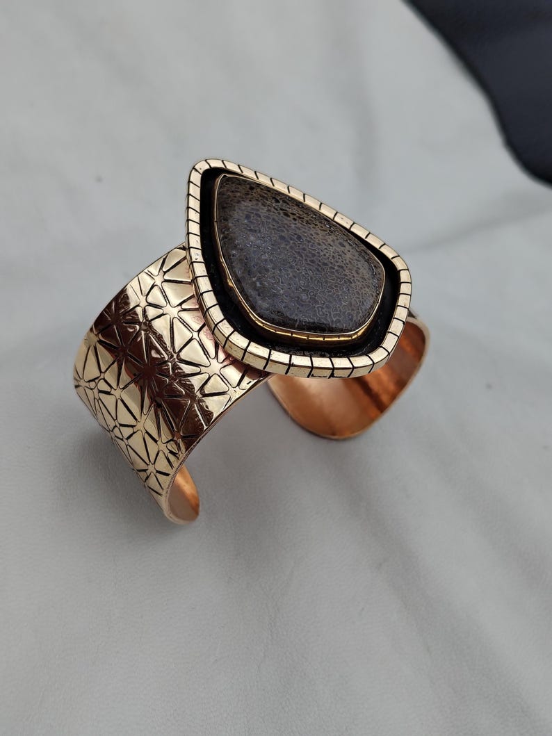 May include: A gold-coloured cuff bracelet featuring a geometric pattern and a teardrop-shaped, dark grey stone. The stone is set in a black and gold frame. The bracelet has a textured surface.