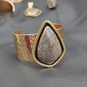 May include: Gold-coloured cuff bracelet with a geometric pattern and a large, teardrop-shaped stone. The stone is framed in gold and black. A matching ring and pendant are visible in the background, suggesting a jewellery set.