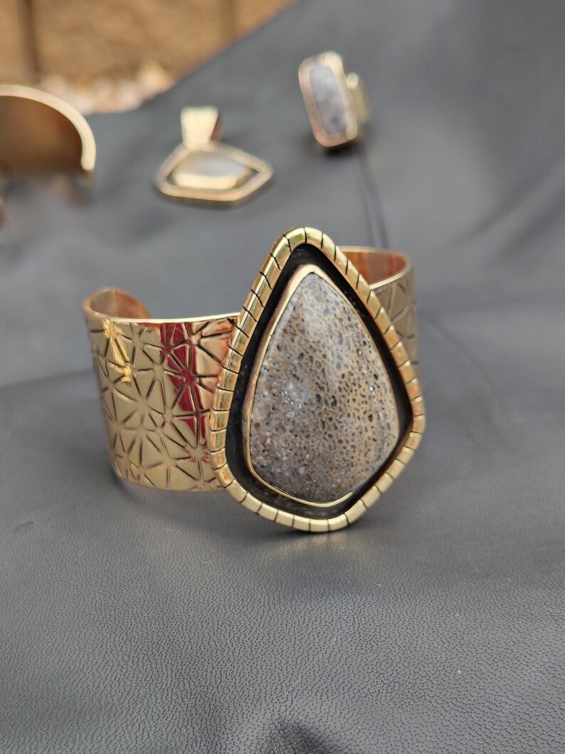 May include: Gold-coloured cuff bracelet with a geometric pattern and a large, teardrop-shaped stone. The stone is framed in gold and black. A matching ring and pendant are visible in the background, suggesting a jewellery set.