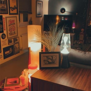 Könnte beinhalten: Ein gemütliches Wohnzimmer mit Retro-Ästhetik. Mit einer Lavalampe, gerahmten Kunstwerken und einem Holzschrank. Eine hohe, zylindrische Lampe und ein kleiner Beistelltisch mit Büchern und Dekoration.