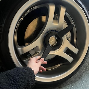 Puede incluir: Llanta de coche plateada y negra con neumático. La llanta tiene un diseño de tres radios con el texto "Super ADVAN Racing" y "ADVAN" en el tapacubos central. La llanta está parcialmente oscurecida por un objeto negro.