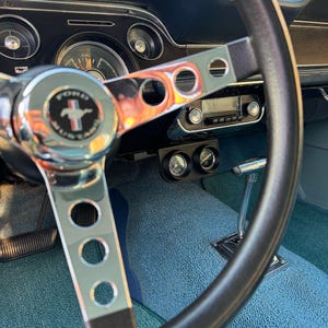 Puede incluir: Primer plano del interior de un coche clásico con un volante negro, una alfombra azul y un salpicadero cromado con indicadores.