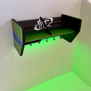 May include: A black and green wall shelf shaped like a car spoiler with a white graphic and five hooks. The shelf has a green light underneath.