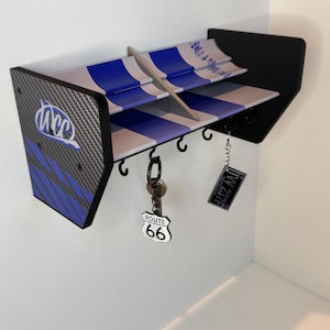 May include: A white and blue racing wing key holder with a carbon fiber pattern and the text "UCC" on the side. The key holder has five hooks and is decorated with a Route 66 keychain and a "Just Married" keychain.